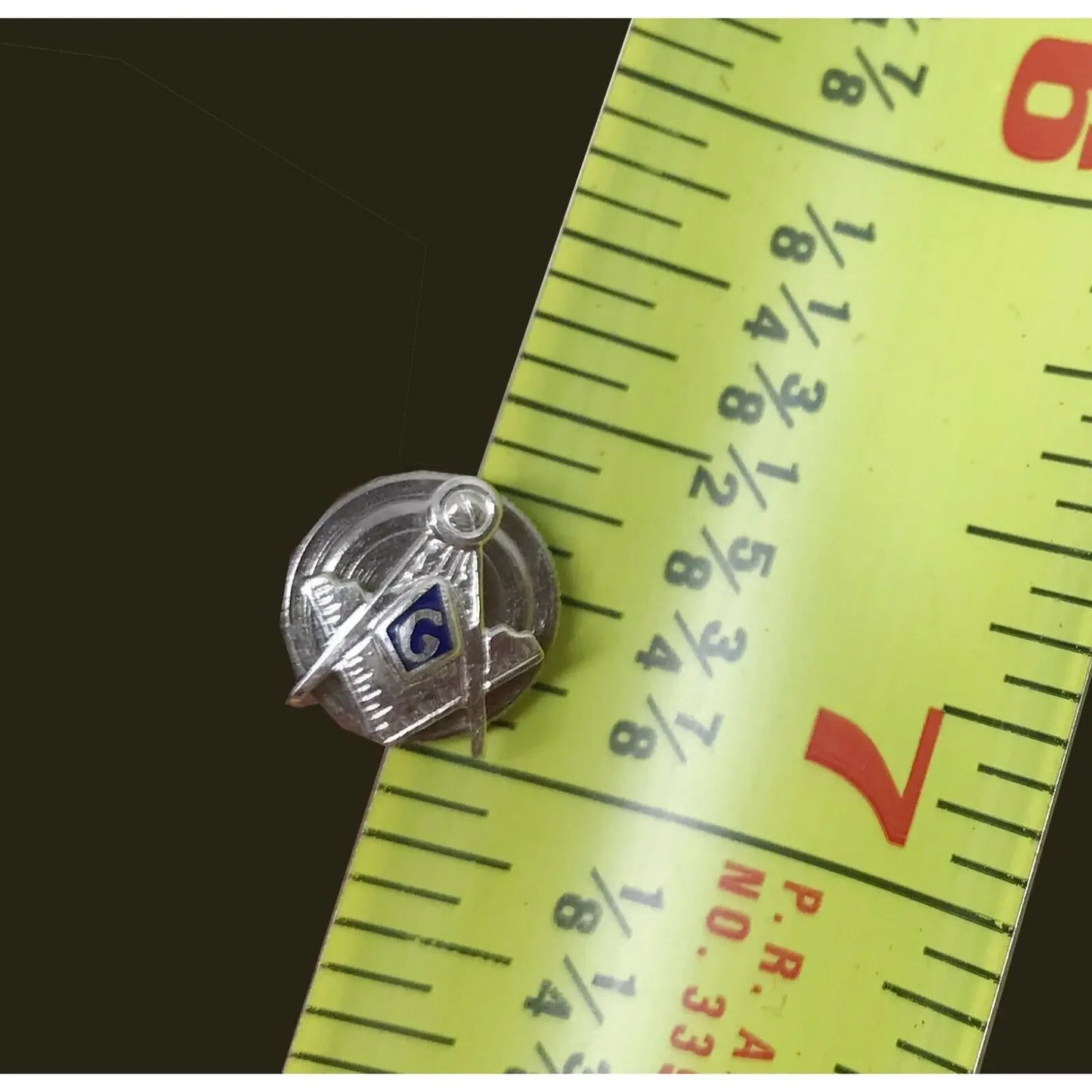Silver Masonic lapel pin with compass and square on yellow measuring tape