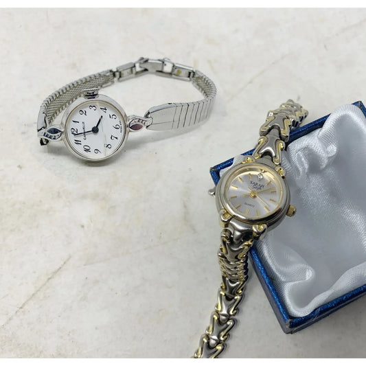 Two women's wrist watches, one silver with a white face and one silver with gold accents in a box, on a light surface.