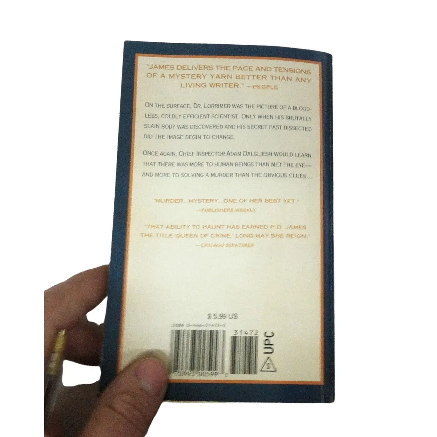 Hand holding a mystery novel with blue cover, back cover text, barcode, and beige background.