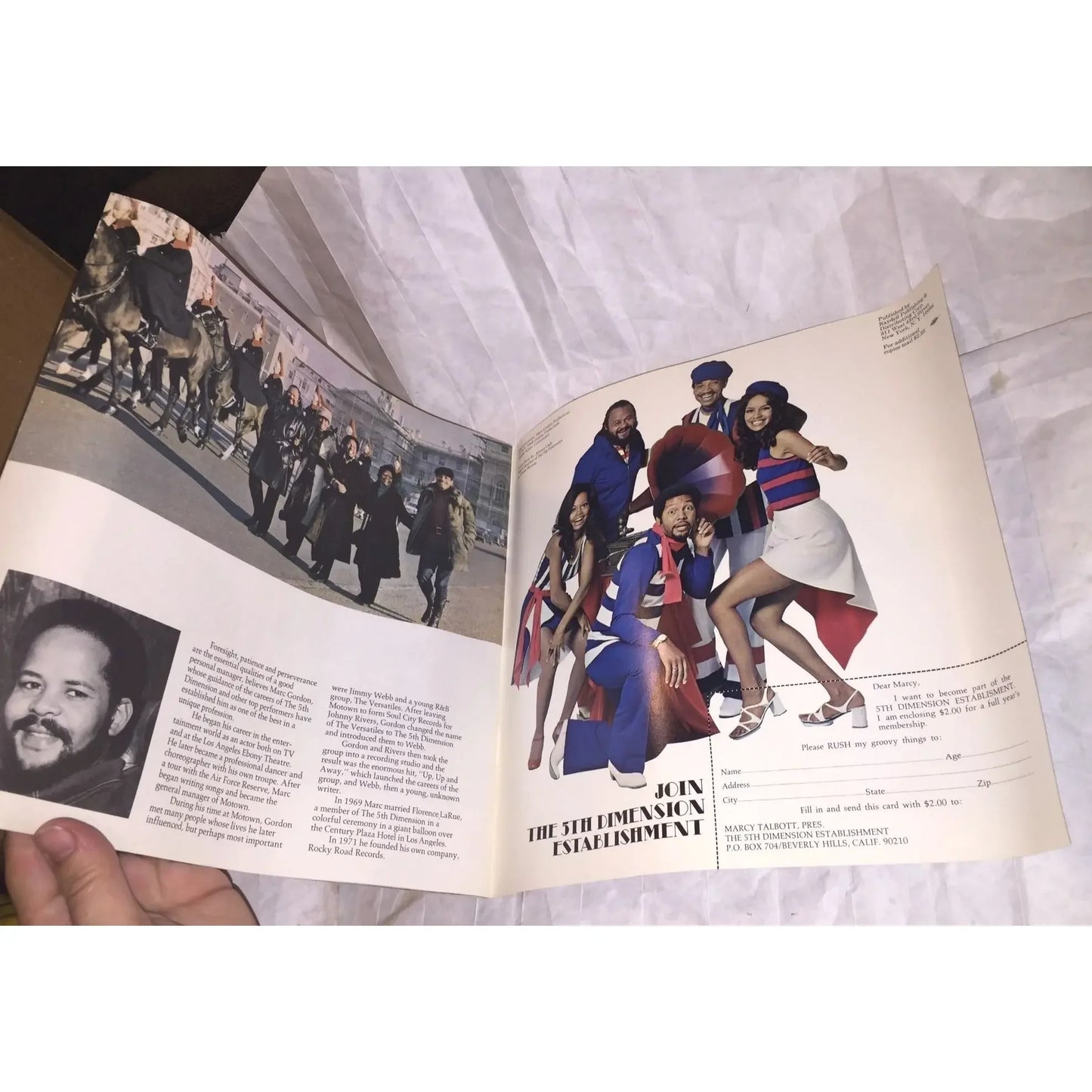 Open magazine held in hand showing a vintage advertisement with a group of people in colorful outfits and a parade scene.